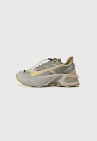 Chaussure de sport grise et beige avec une tige texturée, accents jaunes, bout arrondi et une semelle rembourrée. Présente des lacets élastiques et un logo de marque.