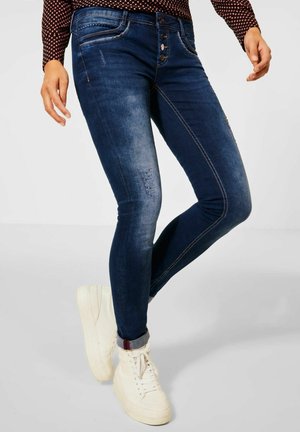 Personne portant un jean skinny bleu foncé avec une fermeture à boutons et des baskets blanches montantes, debout devant un arrière-plan uni.