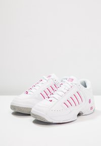 Ett par vita sportskor med rosa ränder och logotyper, grå mönstrade sulor, placerade sida vid sida på en vit yta.