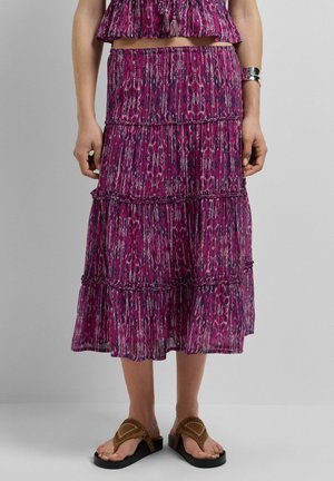 Femme portant une jupe midi à motifs violets et un haut assorti, avec des sandales marron et un bracelet argenté, debout devant un fond gris.