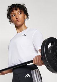 Hvit atletisk T-skjorte med svart logo, kombinert med svarte leggings med grå striper. Personen holder en svart vektskive.