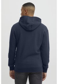 Temno modra trenerka z veliko, strukturirano kapuco, dolgimi rokavi in rebrastimi manšetami. Narejena iz mehkega materiala z gladko teksturo.