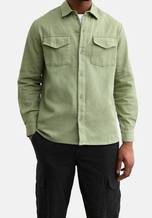 Homme portant une chemise vert clair à boutons avec deux poches poitrine et un pantalon cargo noir, debout avec une main dans la poche.