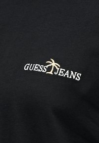 Μαύρο ύφασμα με λευκό κεντητό κείμενο "GUESS JEANS" και ένα μπεζ εικονίδιο φοίνικα ανάμεσα στις λέξεις.