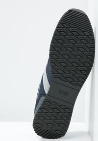 Marinblå atletiska skor med en texturerad tygöverdel, silveraccenter och en mönstrad gummisula med varumärkeslogotypen.