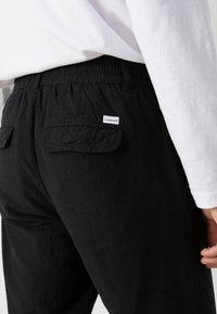 Zwarte broek gemaakt van textiel met structuur, voorzien van een elastische tailleband, twee achterzakken en een klein wit label op de rechterzak.