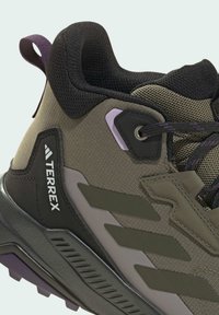 Brown and gray hiking shoe with textured material, prominent logo, eyelet hardware, and purple accents; features a rugged sole and padded collar.