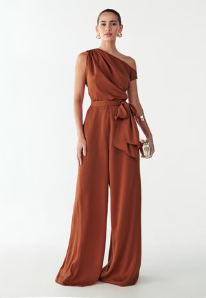 Combinaison marron à une épaule avec ceinture nouée et jambes larges, confectionnée dans un tissu lisse. Comprend un détail drapé prononcé sur l'épaule.