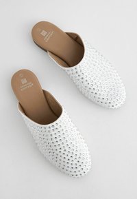 Vita glid-på klackar med en slät yta, prydda med klara rhinestones, med stängd tå och dämpade beige innersulor.