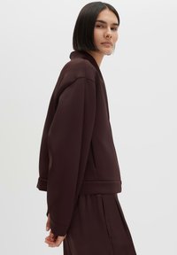Woman with straight dark hair wearing a loose dark brown jacket and matching pants, standing sideways against a plain light background.