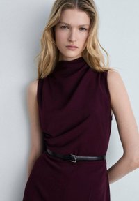 Robe sans manches bourgogne avec un col haut et un design drapé, maintenue par une ceinture noire à la taille, fabriquée à partir d'un tissu doux et texturé.