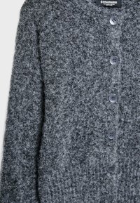 Sivi pleteni kardigan z teksturirano površino, na sprednji strani z temnimi gumbi in sproščenim krojem. Primeren za plastenje v hladnejšem vremenu.