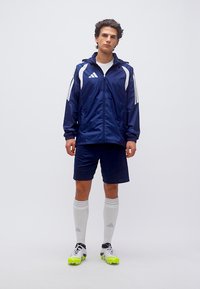 Νέος άνδρας στέκεται φορώντας σκούρο μπλε αθλητικό σακάκι, σκούρα μπλε σορτς, λευκές κάλτσες μέχρι το γόνατο και λευκά ποδοσφαιρικά παπούτσια με νέον κίτρινες σόλες σε απλό φόντο.