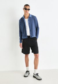 Blue cotton button-up jacket over a white T-shirt, paired with black shorts and gray textured shoes. Model wears dark sunglasses.