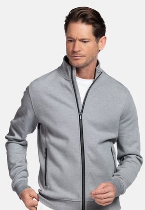 Man draagt lichtgrijs jack met rits over een wit shirt, past de zoom van het jack aan en kijkt iets naar beneden en opzij tegen een effen achtergrond.