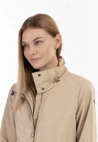Beige vedenpitävä takki korkealla kauluksella, näkyvällä etuvetoketjulla ja napitusnapeilla. Takissa on sileä pinta ja hienovaraisia ompelusaumadetaileja.