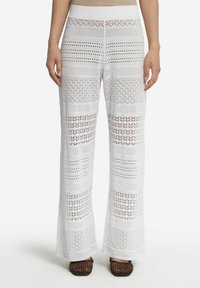 Białe szydełkowe spodnie o szerokim kroju, z teksturowanymi wzorami i otwartymi splotami, zestawione z brązowymi, plecionymi sandałami.