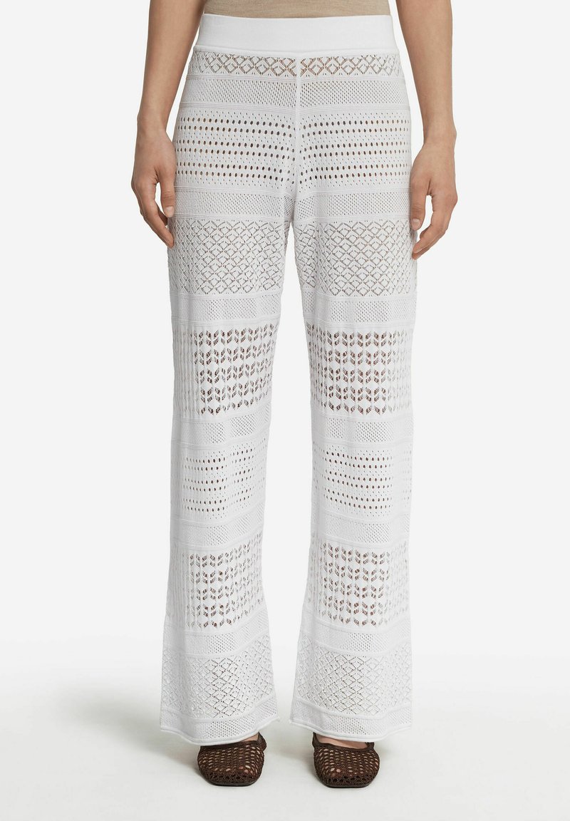 Białe szydełkowe spodnie o szerokim kroju, z teksturowanymi wzorami i otwartymi splotami, zestawione z brązowymi, plecionymi sandałami.