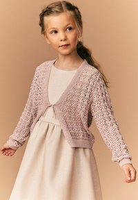 Cardigan tricotat roz deschis, cu design cu dantelă, mâneci lungi și închidere cu fundă, purtat peste o rochie bej strălucitoare.