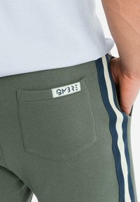 Pantaloni jogger verde oliva con una tasca posteriore, caratterizzati da strisce verticali blu navy e bianche lungo i lati e un'etichetta con il marchio sulla tasca.