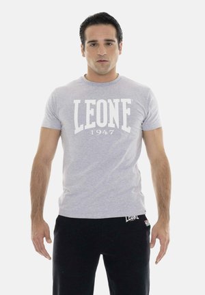 Uomo in piedi di fronte, indossa una maglietta grigio chiaro "LEONE 1947" e pantaloni neri su sfondo bianco.