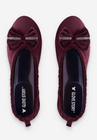Chaussures à enfiler bordeaux avec un nœud en velours et un accent en strass. Design plat avec une bordure texturée et une semelle intérieure rembourrée. Nom de la marque visible.
