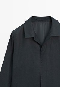 Camicia grigio scuro con colletto, caratterizzata da una texture liscia, una parte anteriore senza bottoni, spalle strutturate e un design pulito e minimalista.