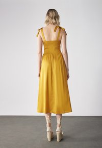 Gul midi-kjole med smock-bodice, justerbare bånd og en flytende skjørt. Kombinert med beige flettet kilehælsandaler.