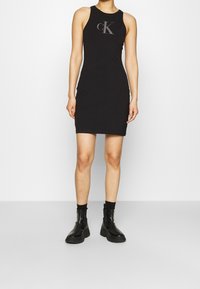 Czarna bawełniana sukienka na ramiączkach o dopasowanym kroju, z błyszczącym logo "CK" na piersi. Do tego czarne botki za kostkę.