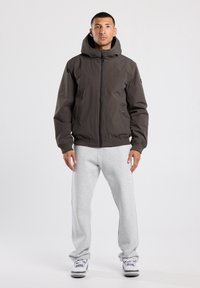 Man standing wearing dark brown hooded jacket, light gray sweatpants, and white sneakers against a plain light background.