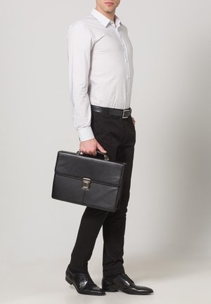 Mies valkoisessa paidassa ja mustissa housuissa, joka pitää mustaa nahkaista salkkua ja seisoo yksinkertaista vaaleanharmaata taustaa vasten.