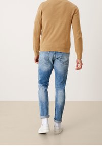 Beżowy sweter, widok z tyłu, w połączeniu z jasnoniebieskimi, lekko przetartymi dżinsami. Białe skarpetki i sneakersy dopełniają stylizację.