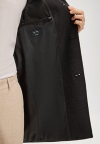 Main tenant une veste en laine noire ouverte, révélant une doublure lisse noire avec une poche intérieure zippée et une étiquette de vêtement.