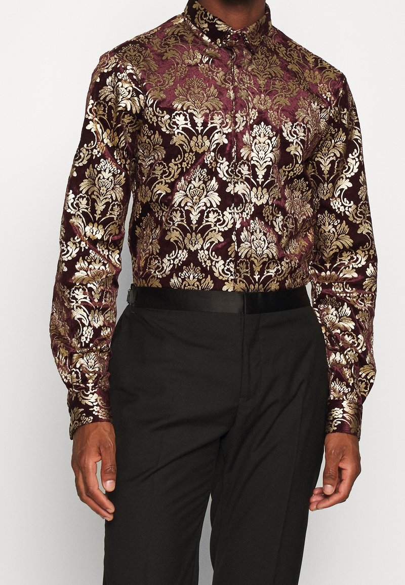 Vínová košile s metalickým zlatým květinovým vzorem, dlouhými rukávy, zapínáním na knoflíky a límečkem, doplněná o jednobarevné černé kalhoty a pásek.