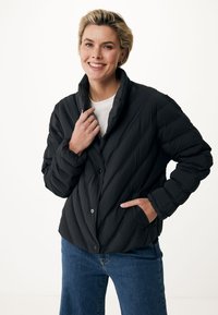 Schwarze, gestepfte Daunenjacke mit hohem Kragen, vorderen Knöpfen und Seitentaschen. Getragen über einem weißen Hemd, mit blauen Jeans.