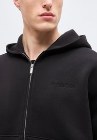 Κοντινή λήψη ενός ατόμου που φοράει μαύρο φούτερ με φερμουάρ και κουκούλα, με το λογότυπο Calvin Klein κεντημένο στο στήθος.