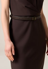 Mørkebrun læderbælte med struktureret overflade, der har en enkel spænde i guldton og flere huller til justering, båret over en kjole.