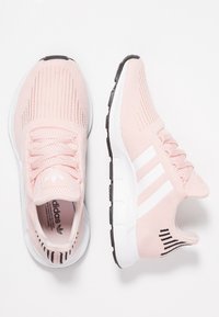 Vaaleanpunaiset urheilukengät, joissa on verkkomainen päällinen, valkoinen kolmiraidallinen logo, teksturoidut yksityiskohdat ja musta kumipohja. Suunniteltu mukavuutta ja joustavuutta ajatellen.