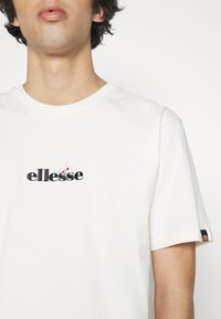 Weißes Baumwoll-T-Shirt mit gesticktem "ellesse"-Logo und floralen Akzenten. Kurze Ärmel, runder Halsausschnitt und ein kleines farbiges Etikett am Ärmel.