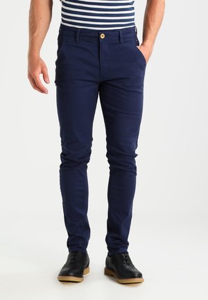 Uomo che indossa pantaloni aderenti blu navy, scarpe di pelle nere e una camicia a righe blu navy e bianche, in piedi con le mani in tasca.