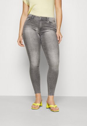 Woman wearing faded gray skinny jeans, a pale yellow top, and bright yellow heeled sandals, standing against a plain light background.