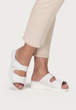 Füße, die weiße Sandalen mit offenen Zehen und verstellbaren Riemen tragen, kombiniert mit hellbeigen, verkürzten Hosen vor einem schlichten Hintergrund.