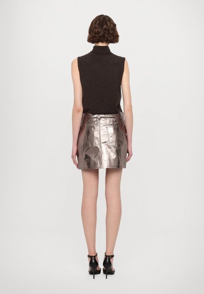 Metalni srebrni mini krilo z žepi, v kombinaciji s temnim brezrokavnikom z visokim ovratnikom. Model nosi črne pete, kar poudarja elegantno silhueto.