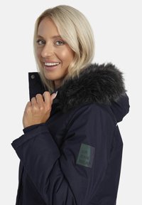 Smaidoša blondīne, kas valkā tumši zilu kapuci jaku ar melnu mākslīgo kažokādu un "Urban Wear" uzšuvi uz piedurknes.