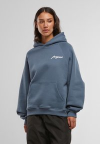 Modrá mikina s kapucňou, predným vreckom, šnúrkou na kapucni a bielym logom na hrudi. Má rebrované manžety a lem, vyrobená z mäkkého materiálu.