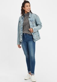 Gaiši zils džinsa jaku, pelēku adītu džemperi, tumši zilus džinsus, baltas kedas. Jakai ir pogu aizdare un nolietots dizains.