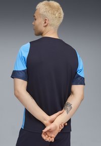 Chemise sportive à manches courtes de couleur marine avec des manches accent bleu clair. Présente un motif texturé bleu sur les poignets des manches. Le matériau est lisse et léger.