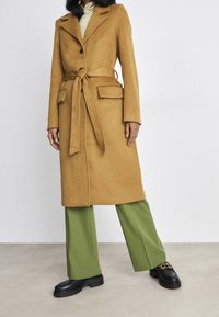 Rock av ullblandning i senapsgult, med midjeband, kavajkrage och framfickor, kombinerad med gröna vida byxor och svarta platåskor.