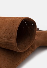 Ruskea mokkanahkasaapas, jossa on ristikkoinen pinta, rento muotoilu ja pehmeä sisus. Varressa on käännetty kukkura ja ommeltu yksityiskohtia.