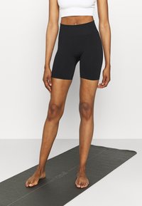 Černé elastické cyklistické šortky s vysokým pasem, hladký materiál, délka po kolena, v kombinaci s bílým přiléhavým krátkým topem. Postava stojí na černé podložce na jógu.